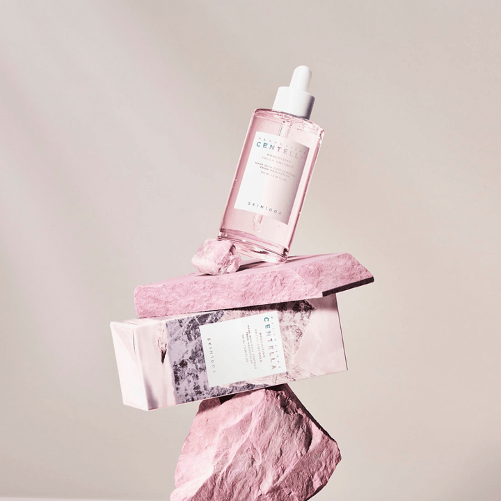 Prozorna steklenička z rožnato ampulo Centella od SKIN1004, uravnotežena na kamnitih blokih, s kartonsko embalažo spodaj, na nevtralni podlagi.