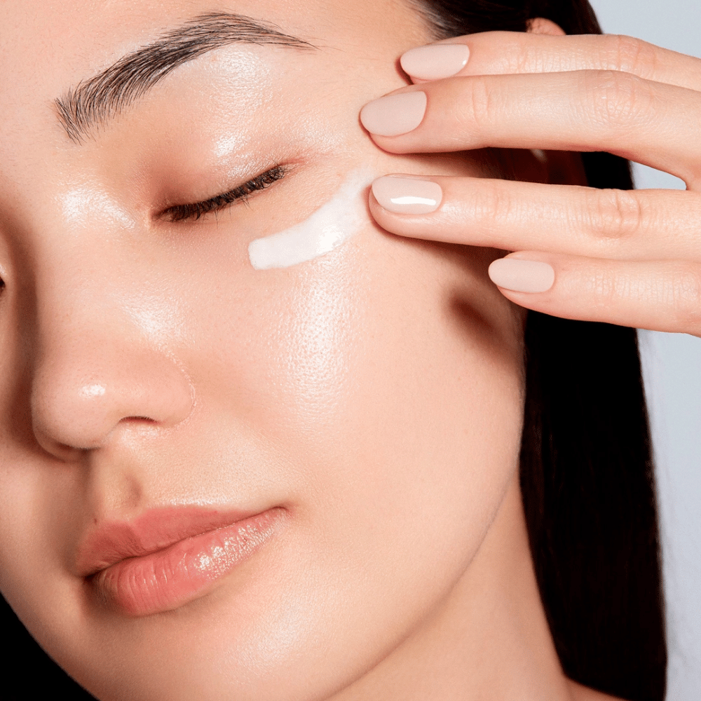 Ženska previdno nanese belo kremo za oči pod oko. Njena koža izgleda sijoča in navlažena, kar poudarja učinek kreme za oči. To izpostavlja izdelek kot vlažilen in primeren za občutljivo kožo okoli oči.