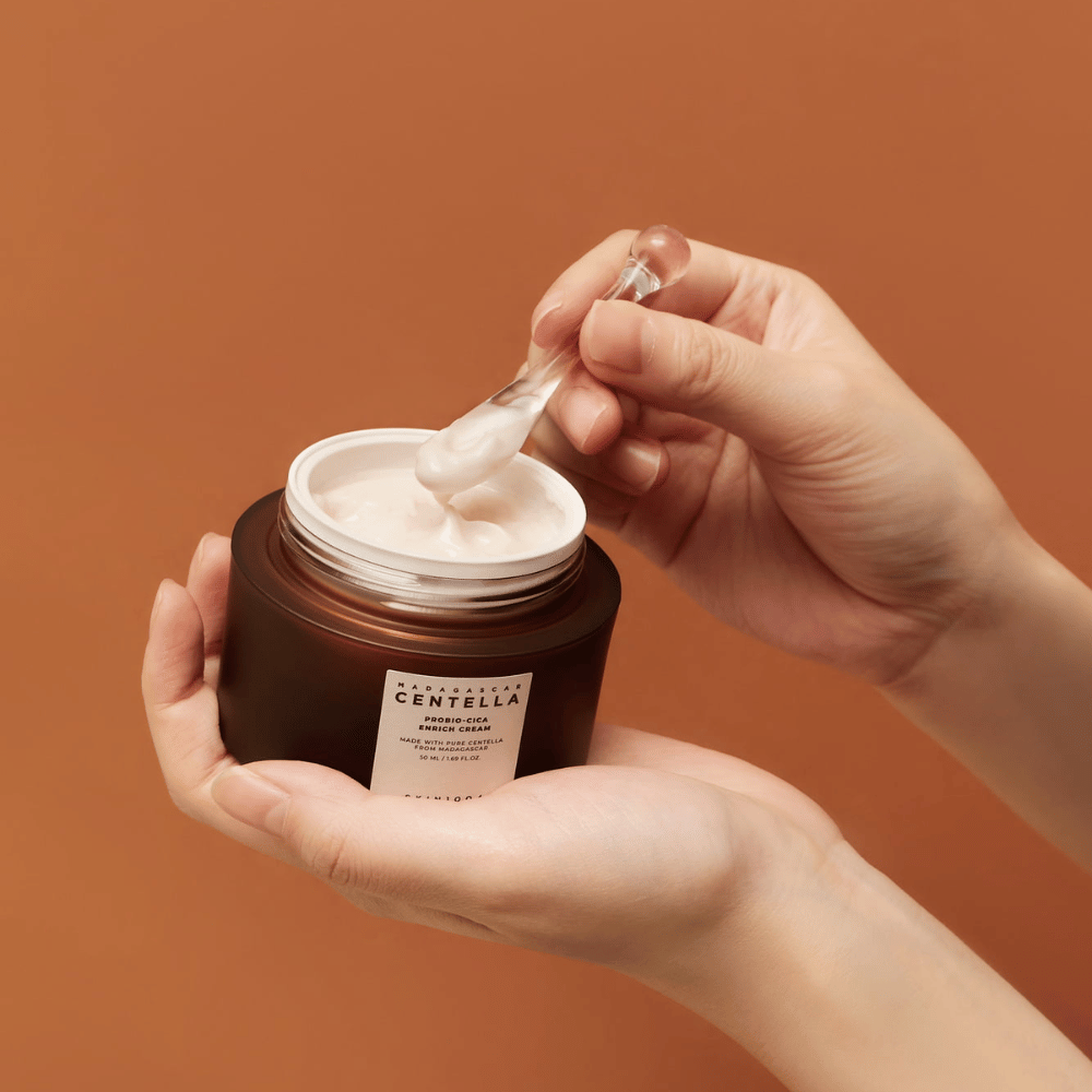 Roke, ki držijo odprto posodo z Madagascar Centella Probio-Cica Enrich Cream in lopatico s kremo na enotnem terakotnem ozadju.