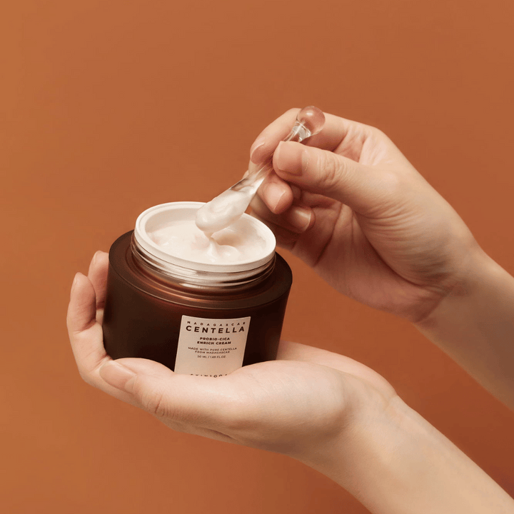 Roke, ki držijo odprto posodo z Madagascar Centella Probio-Cica Enrich Cream in lopatico s kremo na enotnem terakotnem ozadju.