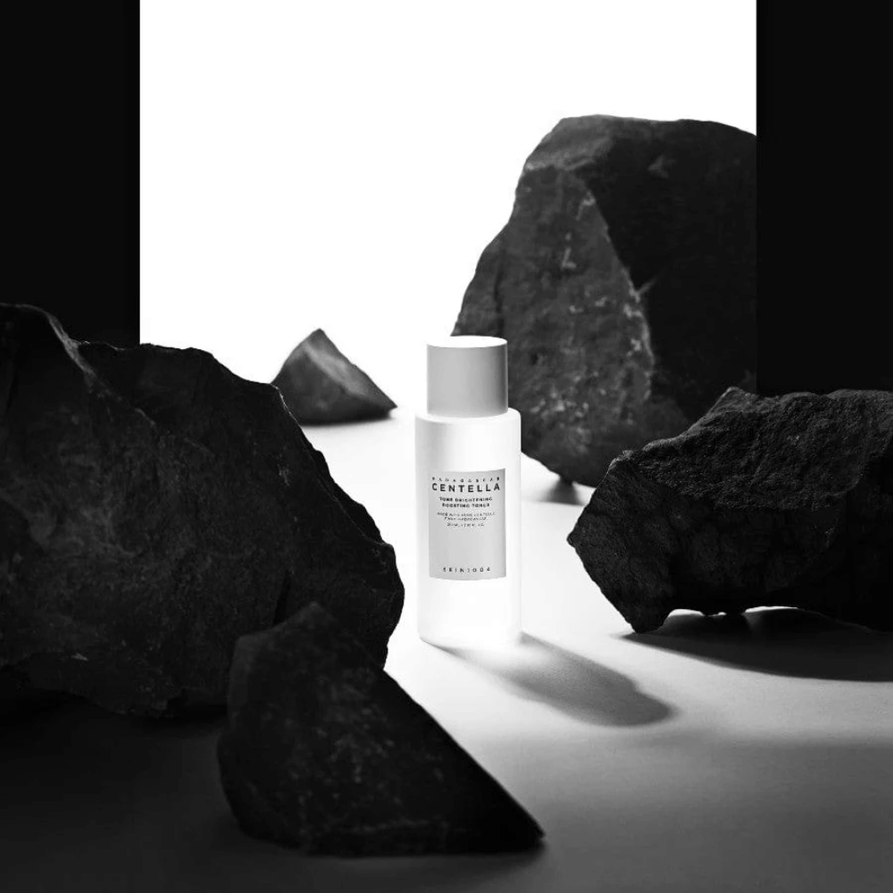 Bela steklenička SKIN1004 Madagascar Centella Tone Brightening Boosting Toner, postavljena dramatično med velike črne kamne na beli podlagi.