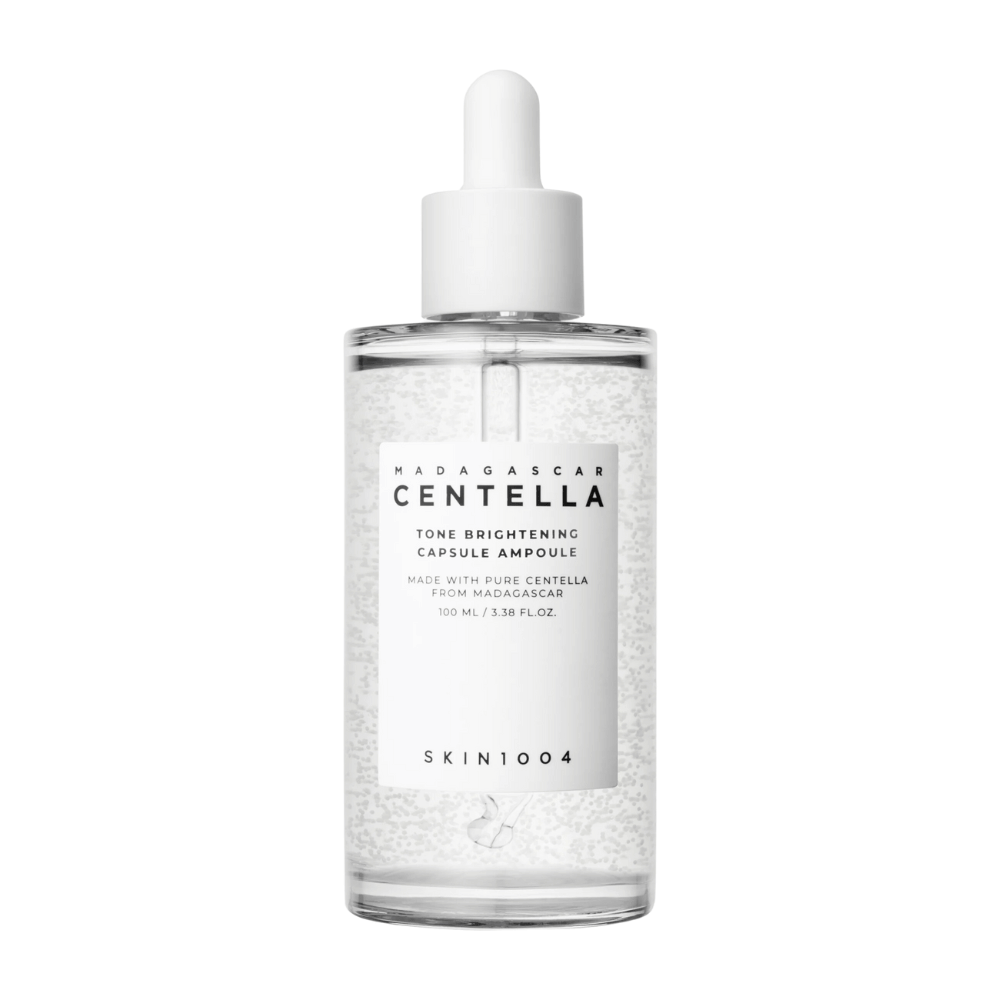 Slika prikazuje stekleničko "SKIN1004 Madagascar Centella Tone Brightening Capsule Ampoule". To je prozorna steklenička z belim dozirnikom. Na etiketi piše, da izdelek vsebuje čiste centella sestavine iz Madagaskarja. Vsebina stekleničke izgleda, kot da vsebuje majhne kapsule ali delce, kar je lahko del njenih svetlečih negovalnih lastnosti. Steklenička vsebuje 100 ml / 3,38 fl.oz. izdelka.
