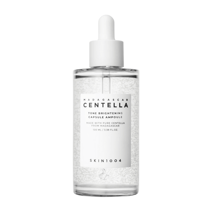 Slika prikazuje stekleničko "SKIN1004 Madagascar Centella Tone Brightening Capsule Ampoule". To je prozorna steklenička z belim dozirnikom. Na etiketi piše, da izdelek vsebuje čiste centella sestavine iz Madagaskarja. Vsebina stekleničke izgleda, kot da vsebuje majhne kapsule ali delce, kar je lahko del njenih svetlečih negovalnih lastnosti. Steklenička vsebuje 100 ml / 3,38 fl.oz. izdelka.