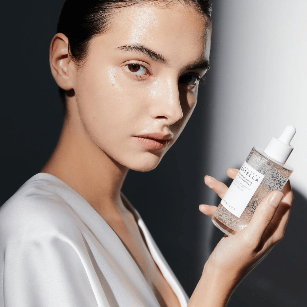 Na sliki je oseba, ki drži stekleničko z dozirnikom. Steklenička je označena z "SKIN1004 Madagascar Centella Tone Brightening Capsule Ampoule". Oseba ima sijoč ton kože, ki odbija svetlobo, kar nakazuje na možen učinek izdelka.