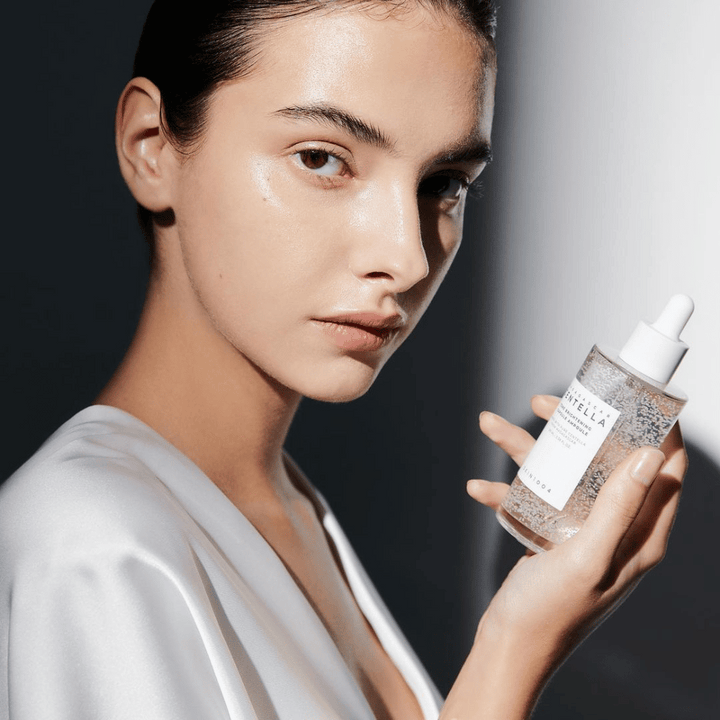 Na sliki je oseba, ki drži stekleničko z dozirnikom. Steklenička je označena z "SKIN1004 Madagascar Centella Tone Brightening Capsule Ampoule". Oseba ima sijoč ton kože, ki odbija svetlobo, kar nakazuje na možen učinek izdelka.