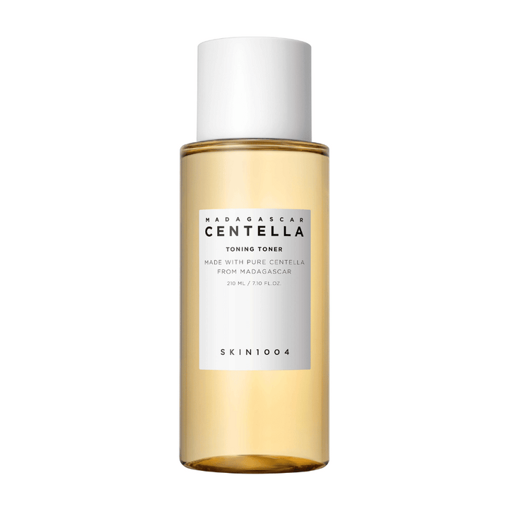 Slika prikazuje steklenico z odtenki za nego kože, označeno "Madagascar Centella Toning Toner" znamke SKIN1004. Izdelek naj bi vseboval 210 ml ali 7.10 fl.oz in je izdelan s čisto centelo iz Madagaskarja. Embalaža je preprosta in elegantna s prozorno steklenico in zlatim toniranim tekočino znotraj, kar daje občutek razkošja in čistosti. Logotip blagovne znamke SKIN1004 je jasno viden v spodnjem delu etikete.
