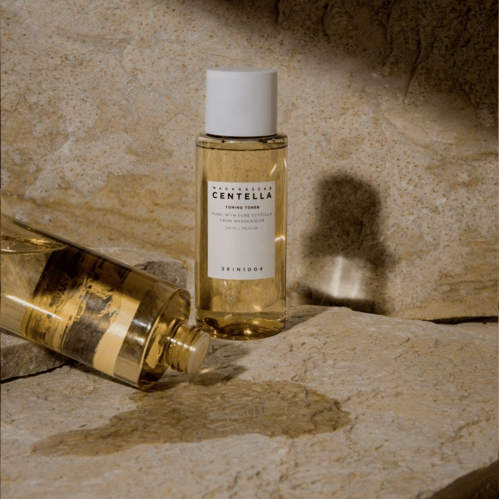 Slika prikazuje steklenico "Madagascar Centella Toning Toner" znamke SKIN1004. Gre za prozorno steklenico, napolnjeno z rumenkasto tekočino, verjetno toner za nego kože. Napisano je, da je narejena s čisto centelo iz Madagaskarja in vsebina je 210 ml / 7.10 fl.oz. Steklenica in ležeča steklenica sta postavljeni na kamnito površino, kar daje naraven in minimalističen občutek.