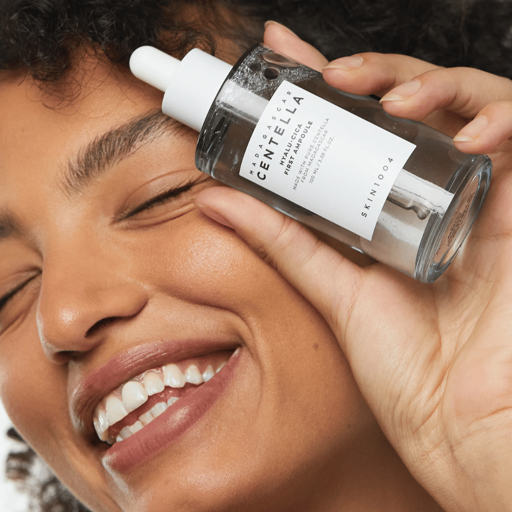 Nasmejana ženska drži Skin1004 Madagascar Centella Hyalu-Cica First Ampoule poleg obraza.