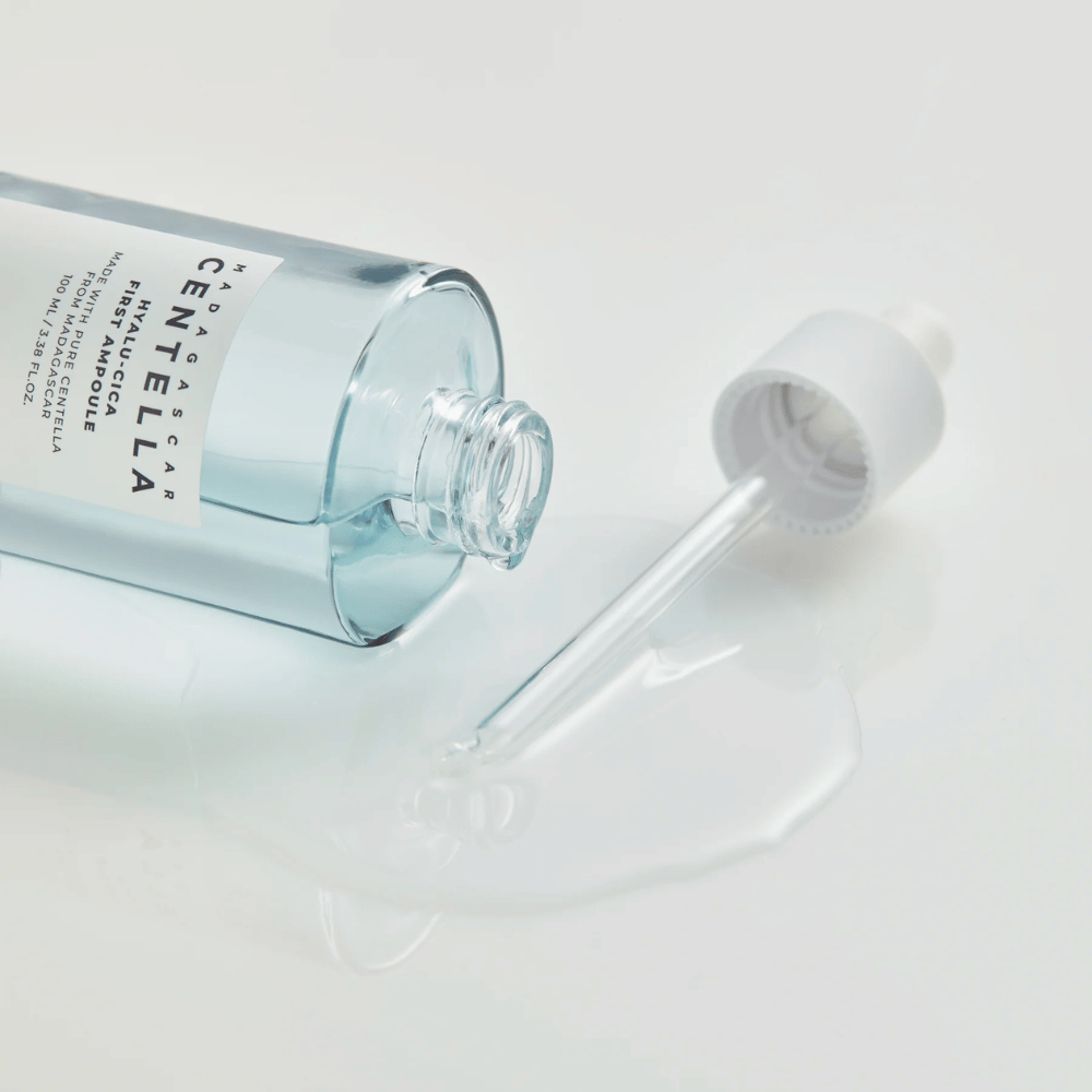 Povečana slika odprte stekleničke Skin1004 Madagascar Centella Hyalu-Cica First Ampoule z ležečo pipeto in serumom razlitim po beli površini.