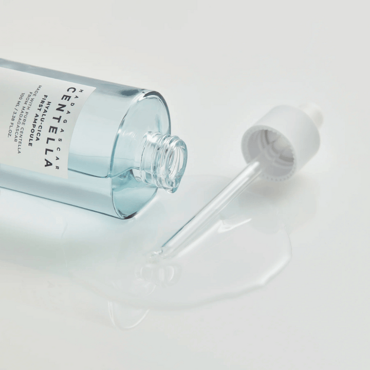 Povečana slika odprte stekleničke Skin1004 Madagascar Centella Hyalu-Cica First Ampoule z ležečo pipeto in serumom razlitim po beli površini.