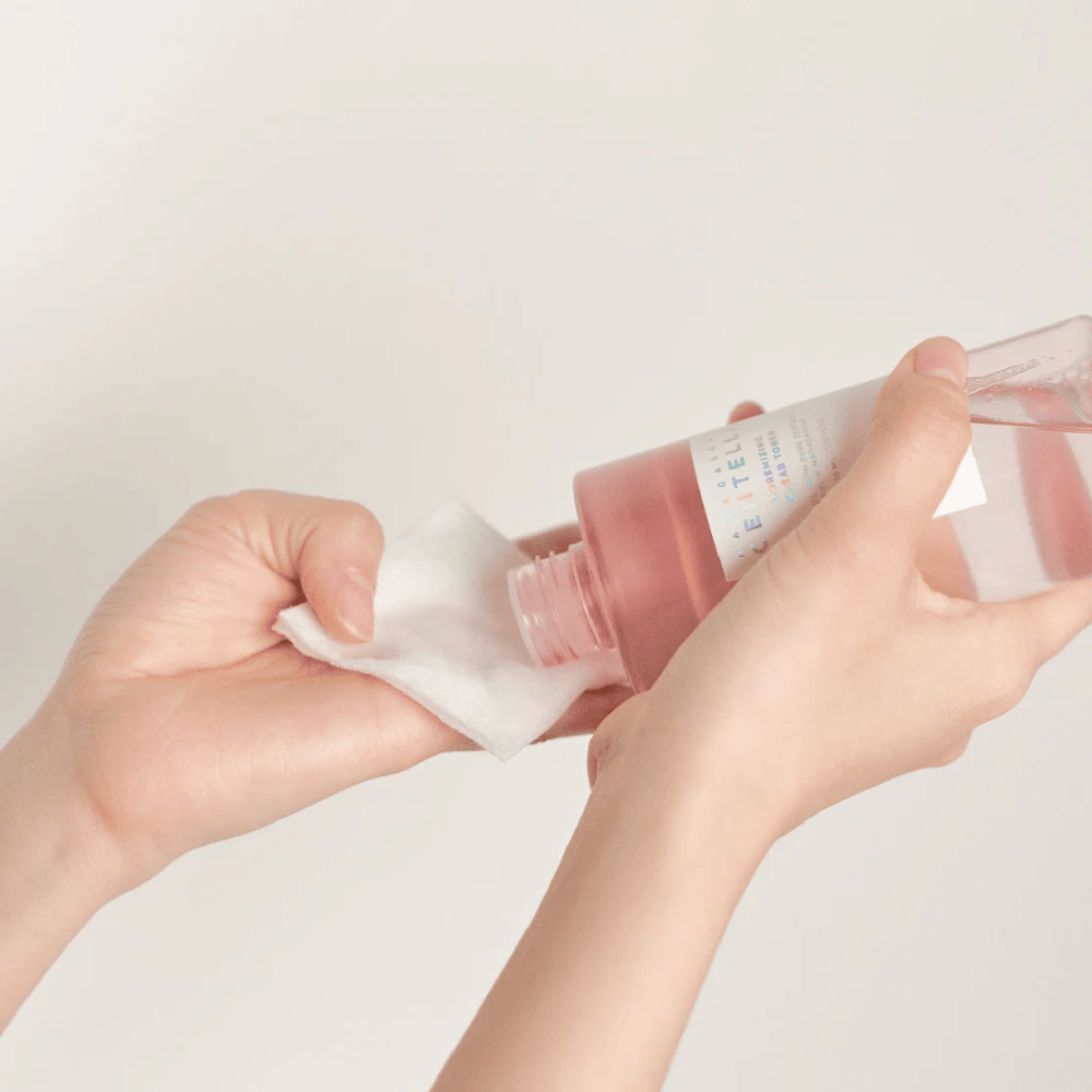 Oseba nalije Madagascar Centella Poremizing Clear Toner iz prozorne steklenice na bombažno blazinico. Izdelek, ki je v roza odtenku, se uporablja za osvežitev in čiščenje kože. Vidne so roke, ki prikazujejo nanos tonika.