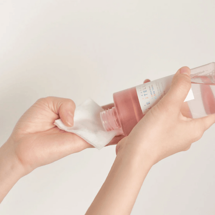 Oseba nalije Madagascar Centella Poremizing Clear Toner iz prozorne steklenice na bombažno blazinico. Izdelek, ki je v roza odtenku, se uporablja za osvežitev in čiščenje kože. Vidne so roke, ki prikazujejo nanos tonika.