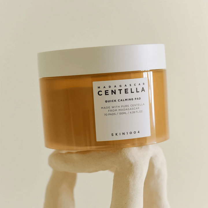 Posoda z SKIN1004 Madagascar Centella Quick Calming Pad postavljena na kiparsko belo podstavek – jantarna posoda z belo etiketo in pokrovom.
