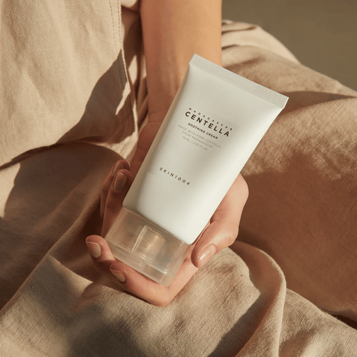 Oseba drži belo tubo Madagascar Centella Soothing Cream od SKIN1004. Tuba ima prozoren pokrovček in besedilo, ki opisuje izdelek kot pomirjujoč in narejen s čisto Centello. Oseba je oblečena v svetla oblačila.