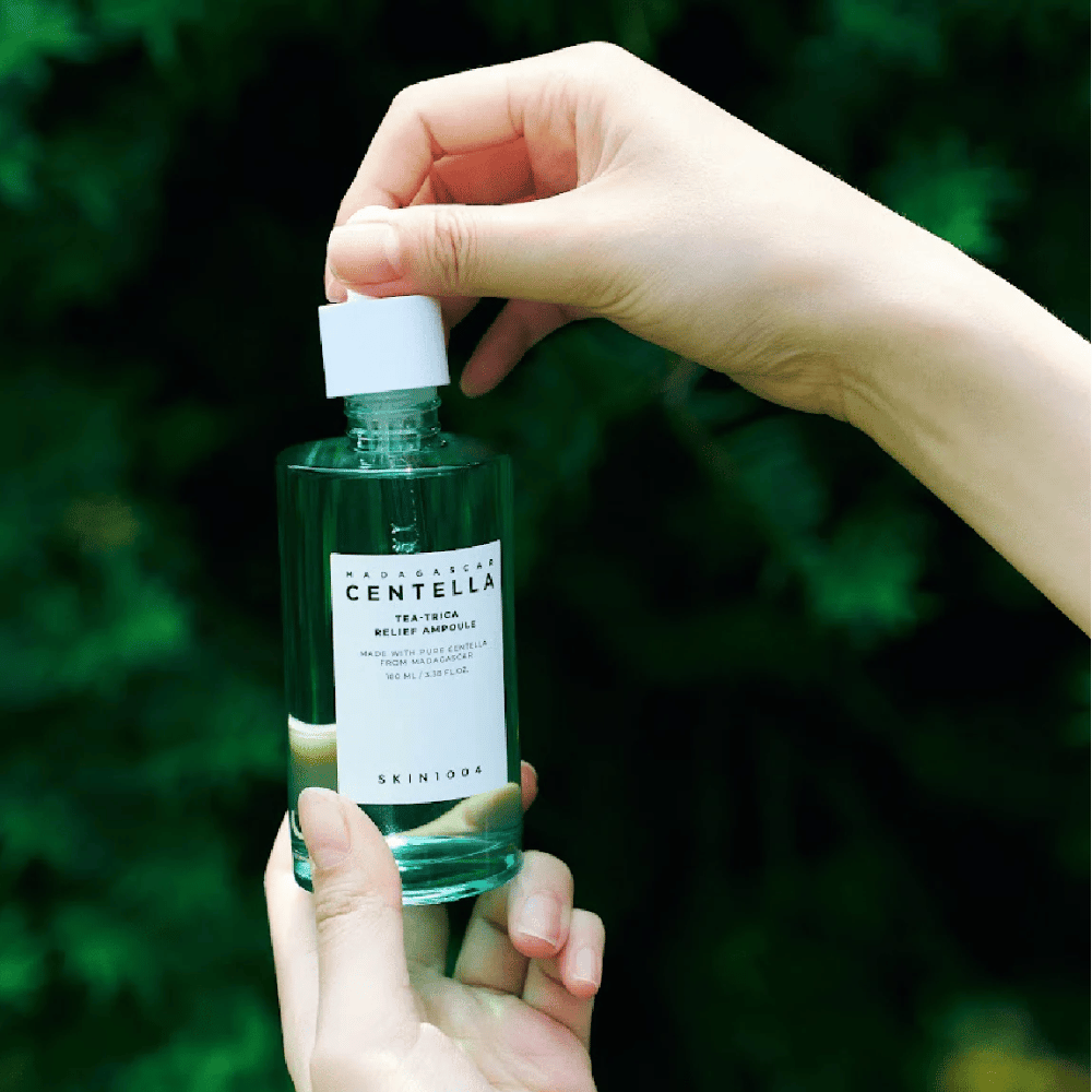 Oseba odpira SKIN1004 Madagascar Centella Tea-Trica Relief Ampoule – zelena steklenica z belo etiketo se drži v rokah pred zelenim ozadjem.