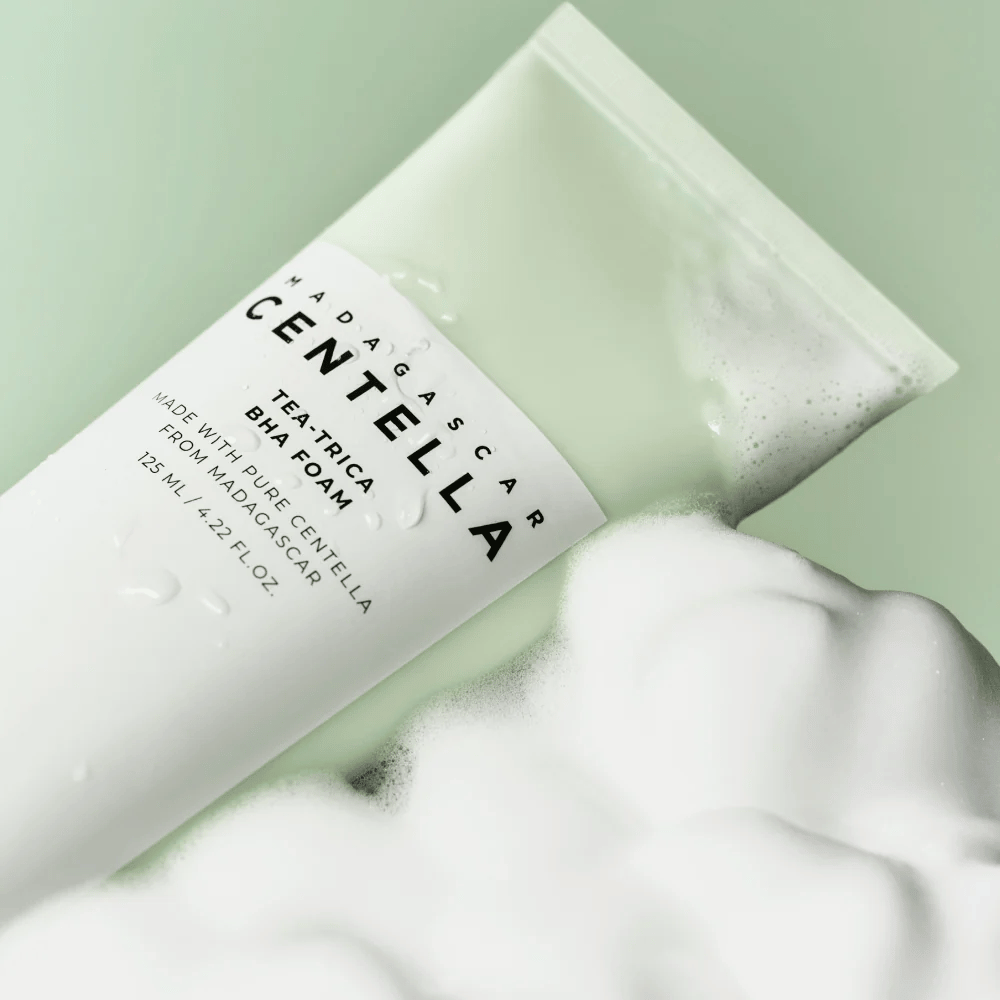 Izdelek tipa čistilna pena, "Madagascar Centella Tea-trica BHA Foam," je delno viden s peno okoli. Izdelek ima zeleno in belo obliko, besedilo "Madagascar Centella" in "Tea-trica BHA Foam" pa sta jasno vidna. Prostornina je 125 ml.