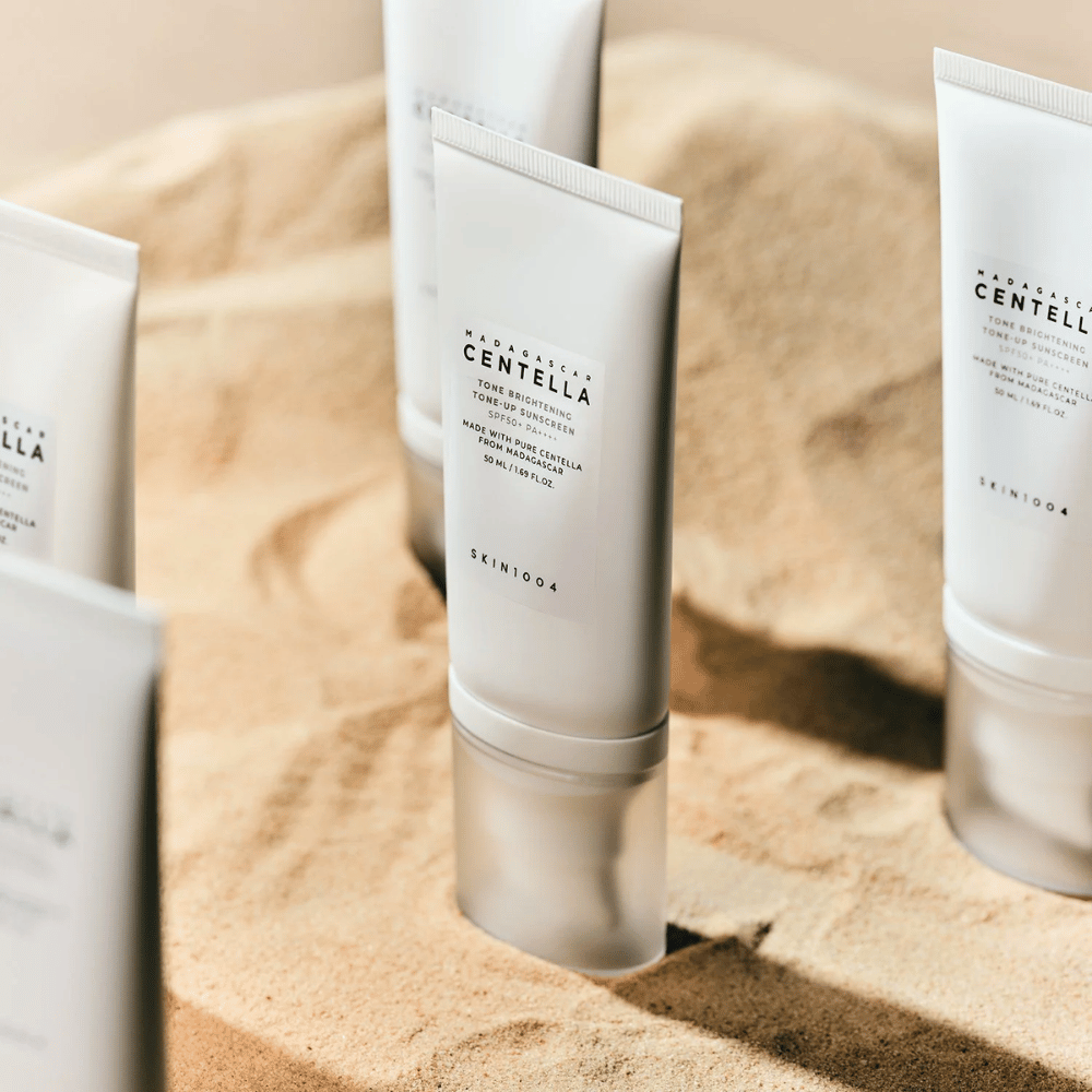 Več tub z Madagascar Centella Tone Brightening Tone-Up Sunscreen stoji v pesku. Tuba je bela s črnim besedilom in vsebuje 50 ml. Izdelek ima SPF 50+ PA++++ in je izdelan s čisto Centello iz Madagaskarja.