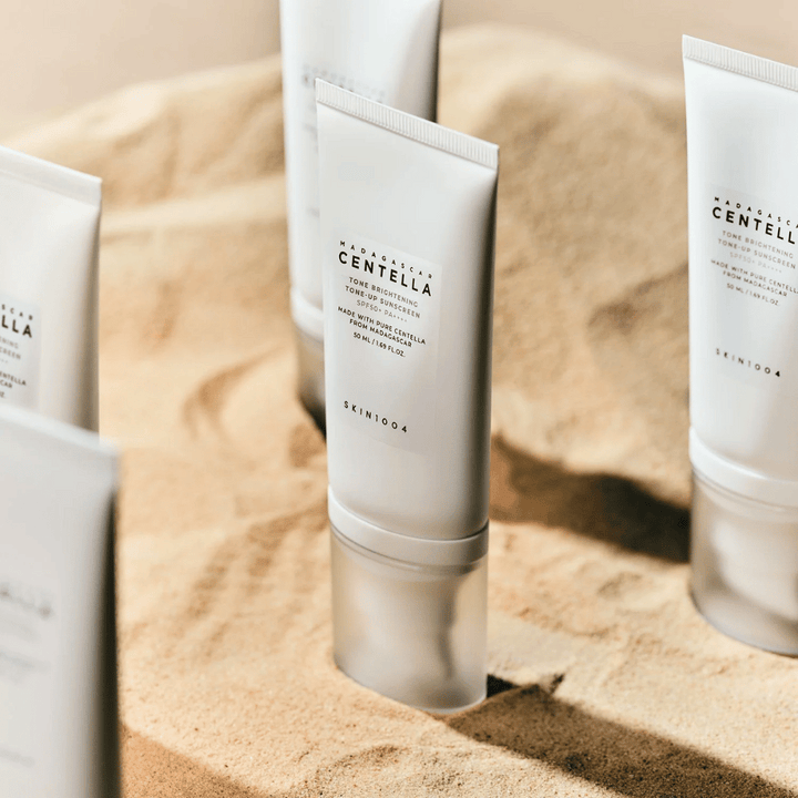 Več tub z Madagascar Centella Tone Brightening Tone-Up Sunscreen stoji v pesku. Tuba je bela s črnim besedilom in vsebuje 50 ml. Izdelek ima SPF 50+ PA++++ in je izdelan s čisto Centello iz Madagaskarja.