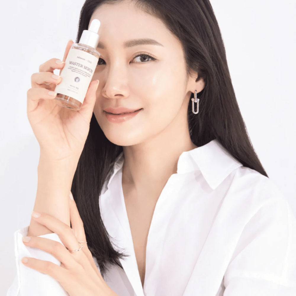 Nasmejana ženska z dolgimi, temnimi lasmi in belo srajco drži Mixsoon Master Serum poleg svojega obraza. Njena koža izgleda sijoča in navlažena.
