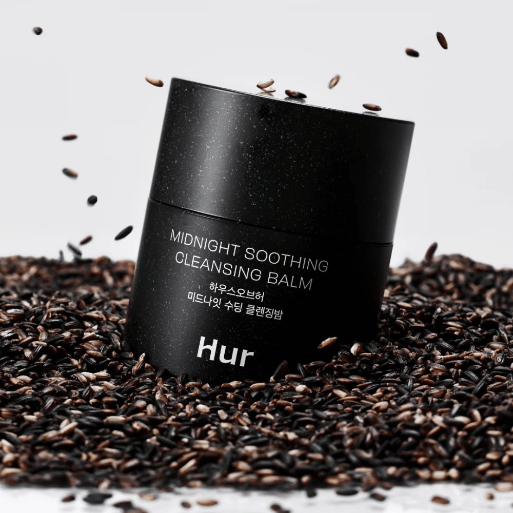 Kako Midnight Soothing Cleansing Balm v črni posodi postavljen na črna semena. Pomirjujoč čistilni balzam, ki raztaplja ličila in nečistoče, obenem pa neguje kožo.