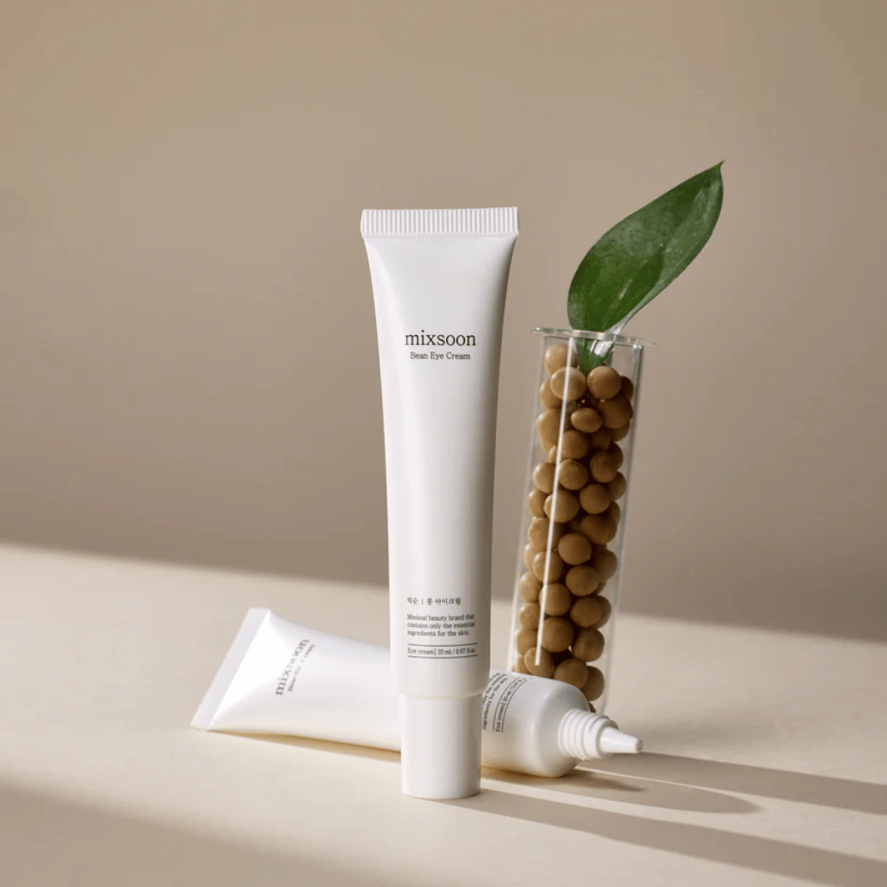 Dve tubi Mixsoon Bean Eye Cream postavljeni poleg epruvete, napolnjene s sojinimi zrni, in zelenim listom rastline, na bež površini s sencami.