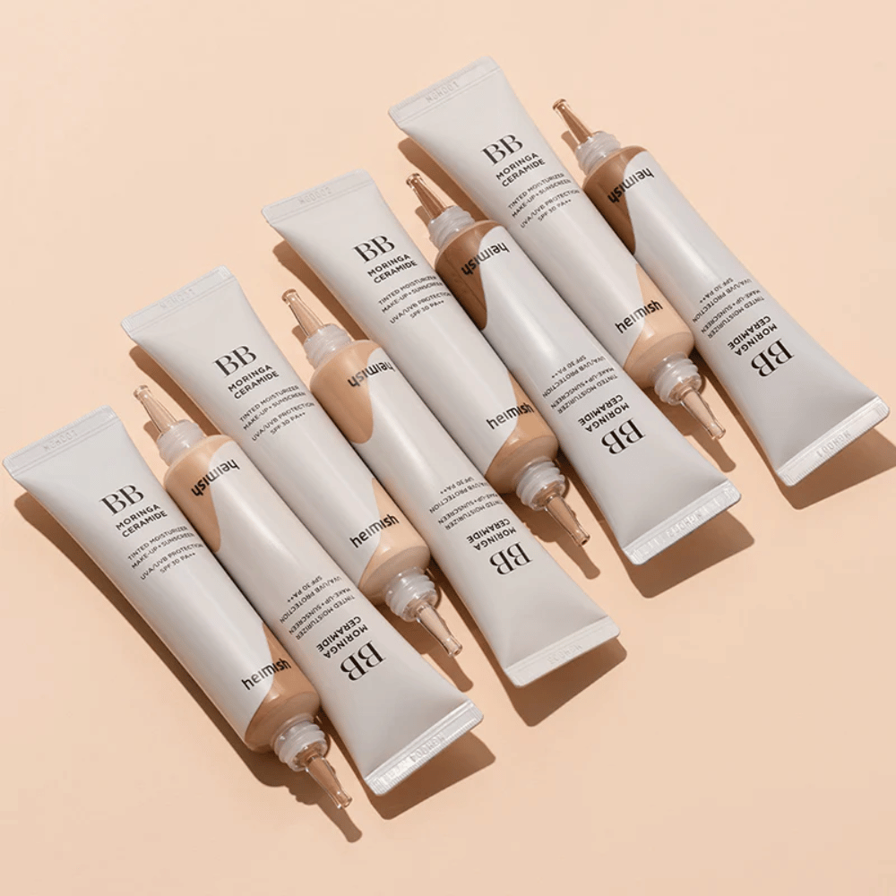Več tub Heimish Moringa Ceramide BB Cream razporejenih na svetlo bež ozadju. Slika poudarja različne odtenke in elegantno, minimalistično zasnovo embalaže izdelka.