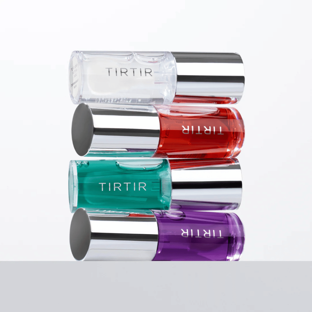 Štiri TIRTIR My Glow Lip Oil v različnih barvah – prozorna, rdeča, zelena in vijolična – zložene ena na drugo s srebrnimi pokrovčki na beli ozadju.