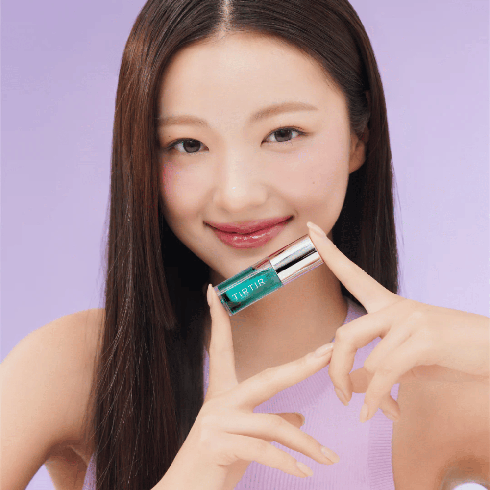 Ženska z dolgimi, ravnimi lasmi drži TIRTIR My Glow Lip Oil in razkazuje svoje sijoče, rožnate ustnice proti vijoličnemu ozadju.