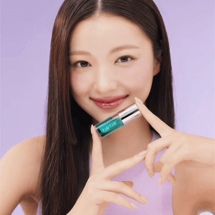 Ženska z dolgimi, ravnimi lasmi drži TIRTIR My Glow Lip Oil in razkazuje svoje sijoče, rožnate ustnice proti vijoličnemu ozadju.