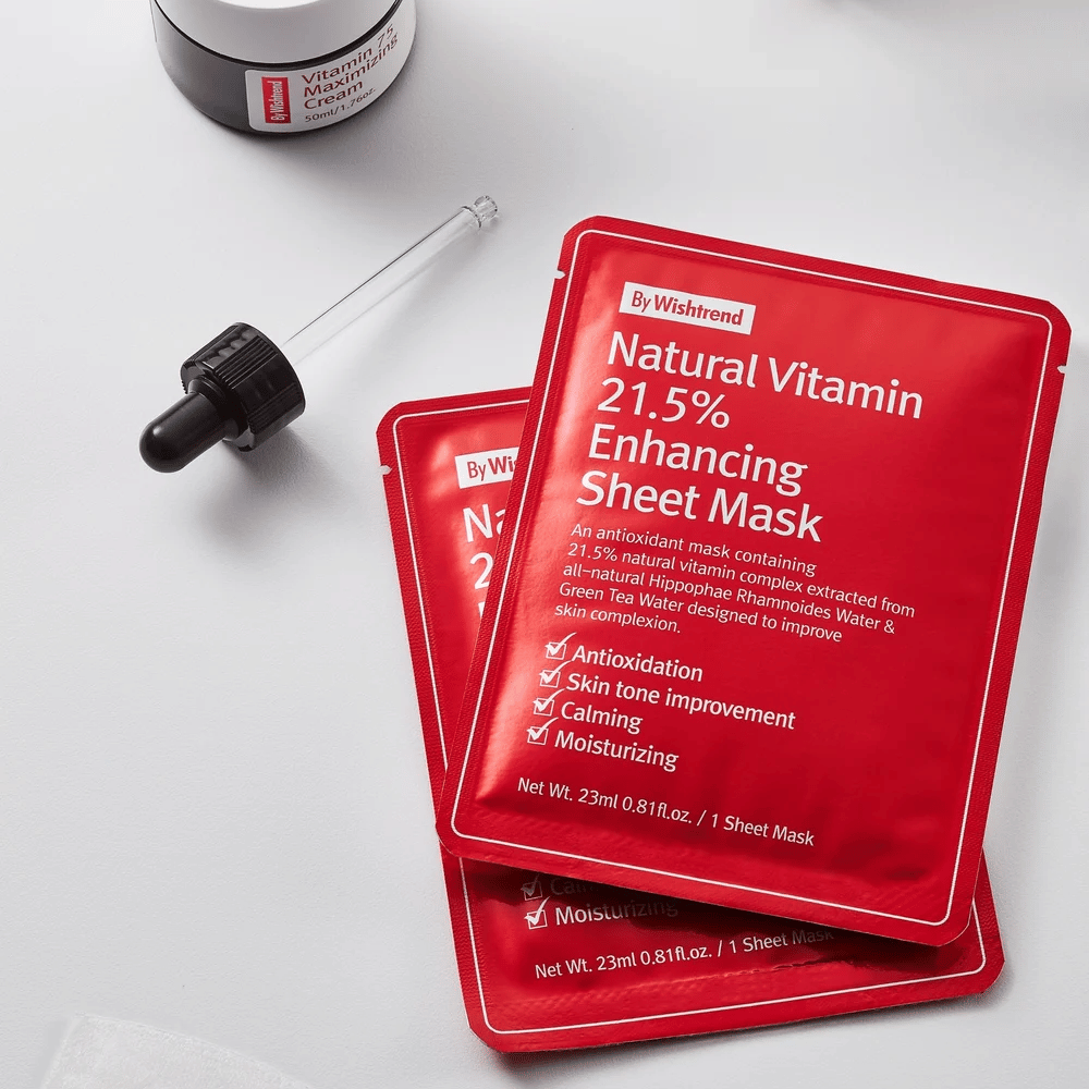 By Wishtrend Natural Vitamin 21.5% Enhancing Sheet Mask v rdečih embalažah, postavljenih na belo površino skupaj s pipeto in lončkom za obrazno kremo.