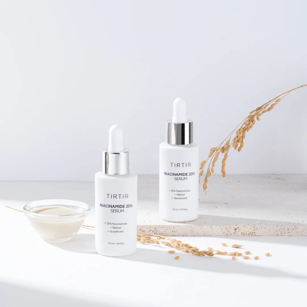 Dve steklenički Tirtirs "Niacinamide 20% Serum" sta prikazani na svetli površini skupaj z majhno skledo s kremasto snovjo in nekaj suhimi rastlinskimi vejicami. Serum vsebuje 20 % niacinamida, retinola in glutationa, kar prispeva k izboljšanju videza in teksture kože.