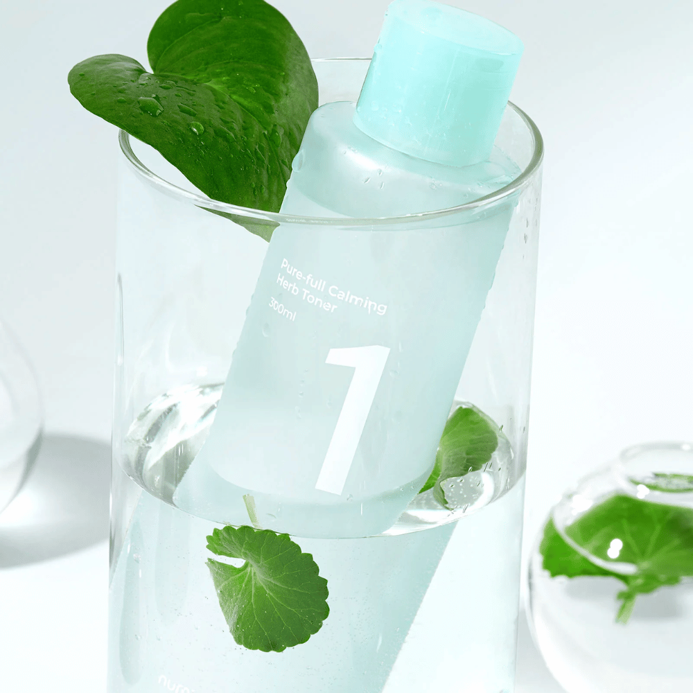 Steklenica No.1 Pure-Full Calming Herb Toner postavljena v kozarec z vodo in zelenimi listi. Steklenica ima svetlo moder pokrov in etiketo z napisom "Pure-full Calming Herb Toner" ter "300ml". Izdelek izgleda sveže z vodo in rastlinjem v ozadju, kar daje pomirjujoč in naraven občutek.