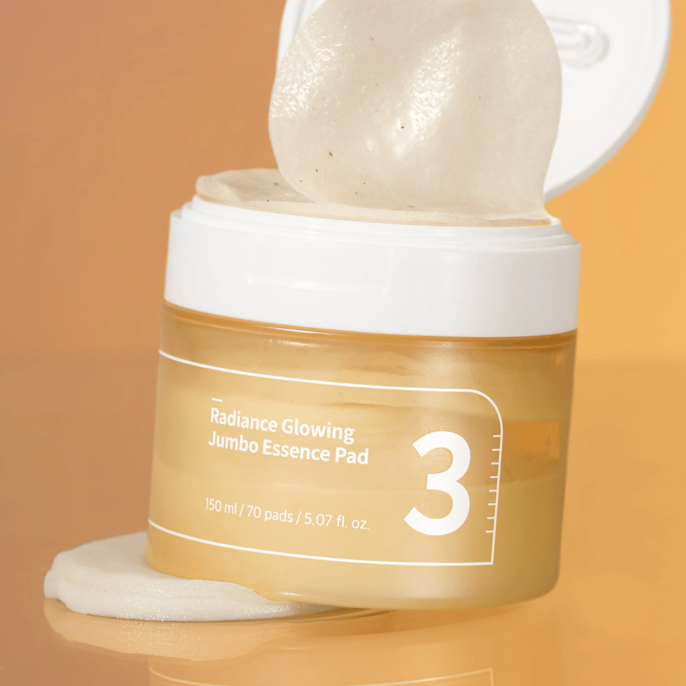 Odprt kozarec z numbuzin No. 3 Radiance Glowing Jumbo Essence Pad, ena blazinica je nagnjena proti robu, ozadje v oranžnem tonu.