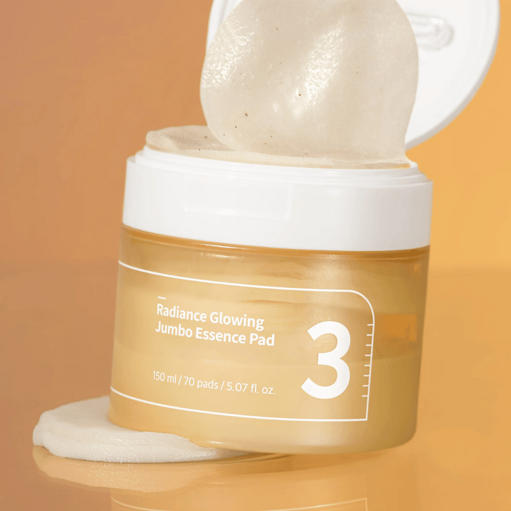 Odprt kozarec z numbuzin No. 3 Radiance Glowing Jumbo Essence Pad, ena blazinica je nagnjena proti robu, ozadje v oranžnem tonu.