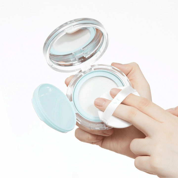 Roka drži odprto posodo z VT PDRN Essence Glow Sun Pact. Blazinica je na poti, da nanese puder, ki ima belo površino in moder rob okoli posode.