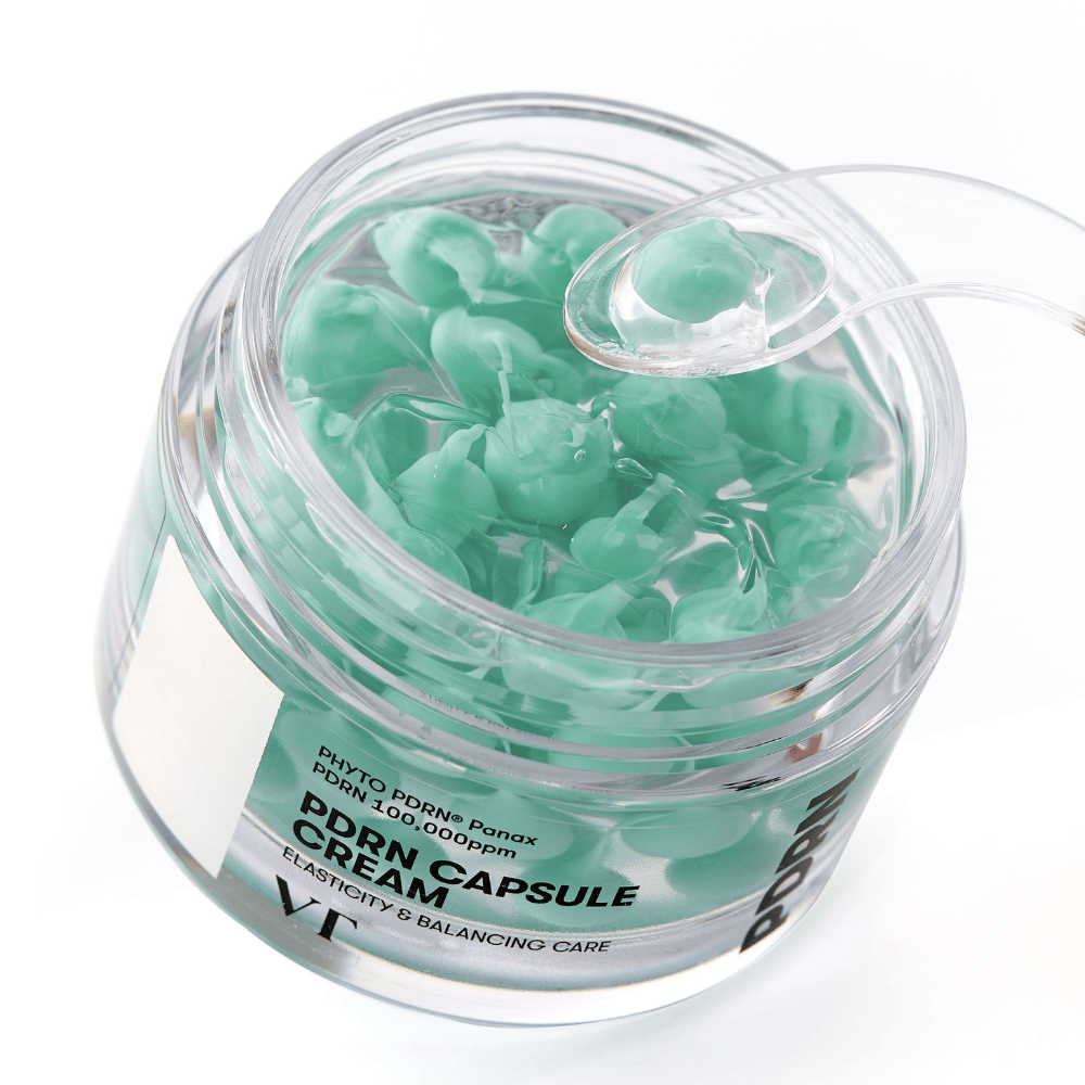 Približan posnetek odprte posode z VT PDRN Capsule Cream. Vsebina je sestavljena iz majhnih zelenih kapsul v prozorni gelu, s priloženo lopatico za nanašanje. Besedilo na posodi se glasi "Elasticity & Balancing Care".