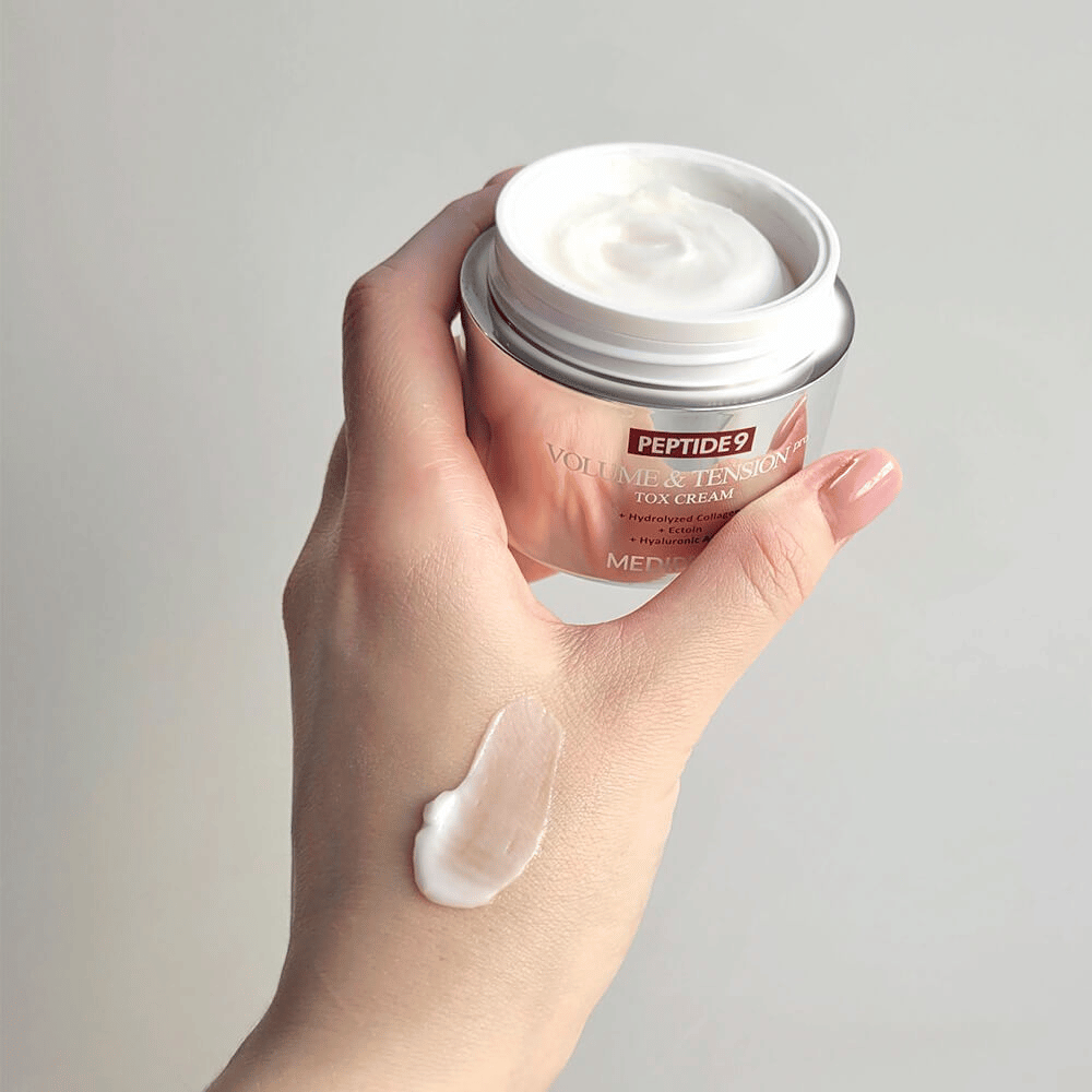 Roka drži MEDI-PEEL Peptide 9 Volume & Tension Tox Cream s kančkom kreme na hrbtni strani roke. Svilnata formula vlaži in daje koži čvrstost in prožnost.