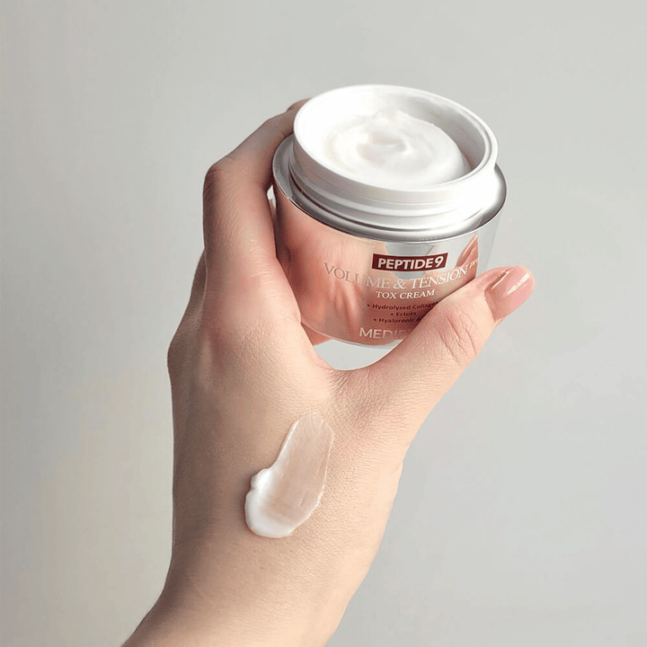 Roka drži MEDI-PEEL Peptide 9 Volume & Tension Tox Cream s kančkom kreme na hrbtni strani roke. Svilnata formula vlaži in daje koži čvrstost in prožnost.