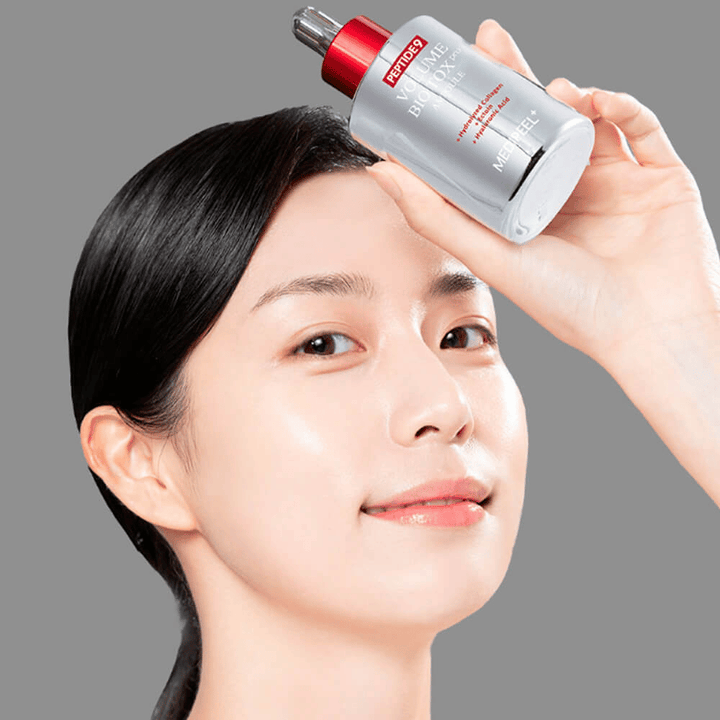 Oseba drži MEDI-PEEL Peptide 9 Volume Bio Tox Pro Ampoule proti čelu. Koža izgleda navlažena in sijoča – serum krepi elastičnost in čvrstost.