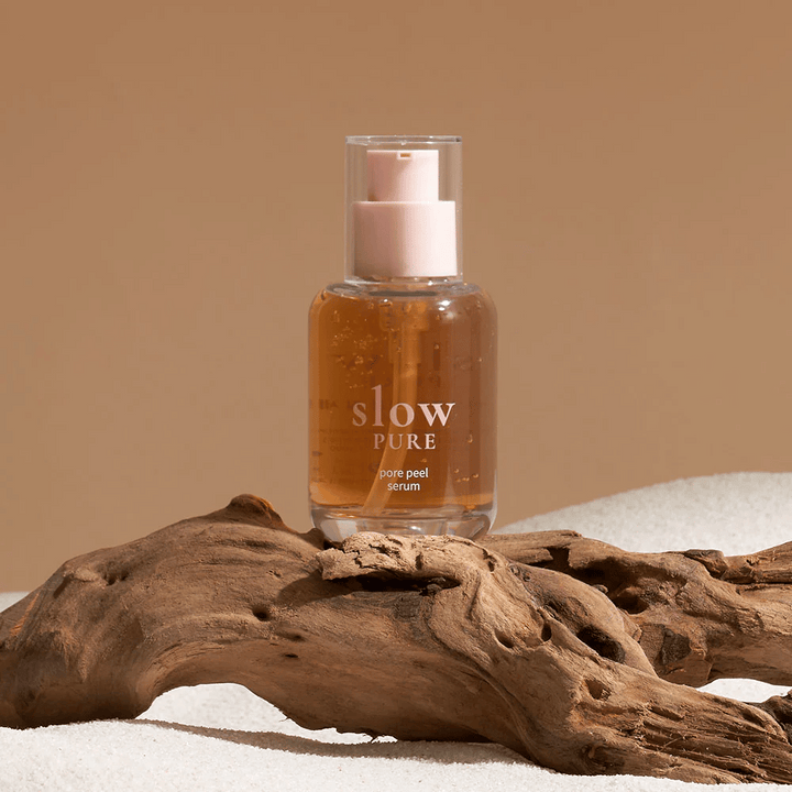 Slow Pure Pore Peel Serum postavljen na pesek z leseno podrobnostjo v ozadju, simbol za naravno nego kože.