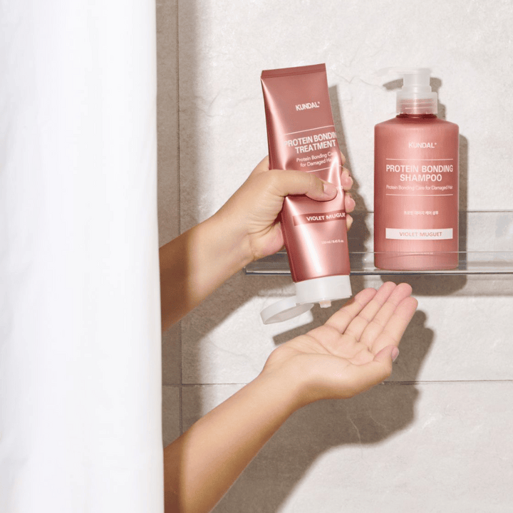 KUNDAL Protein Bonding Treatment Violet Muguet se uporablja pod prho, s Protein Bonding Shampoo postavljenim na polico poleg.