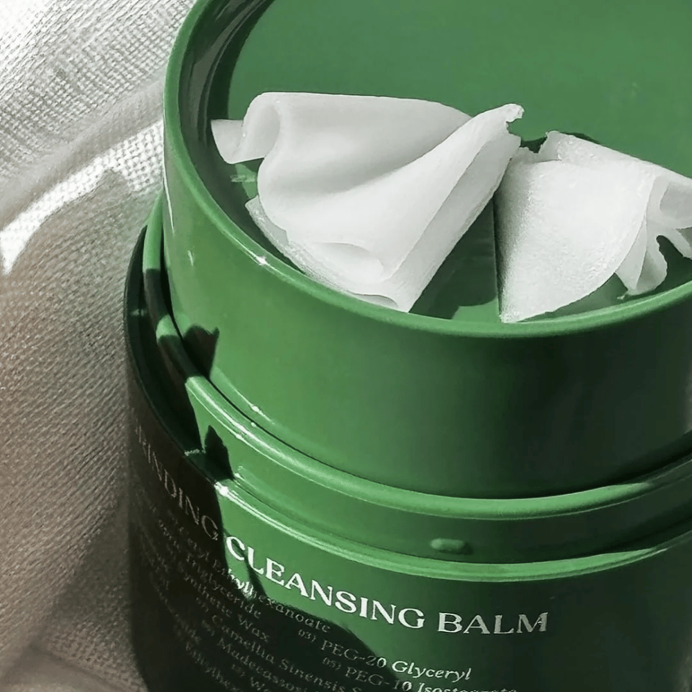 Slika prikazuje zeleno posodico z "Cleansing Balm", ki je odprta, da razkrije priloženo lopatico in mehko vsebino. Kremasta tekstura odbija svetlobo, kar daje občutek razkošja in učinkovitosti pri čiščenju.