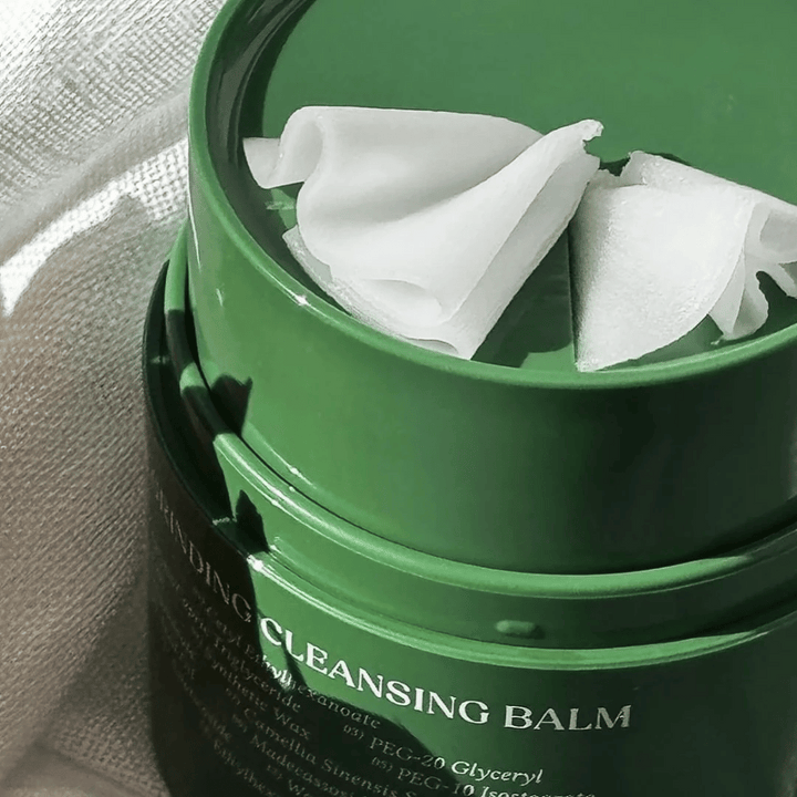 Slika prikazuje zeleno posodico z "Cleansing Balm", ki je odprta, da razkrije priloženo lopatico in mehko vsebino. Kremasta tekstura odbija svetlobo, kar daje občutek razkošja in učinkovitosti pri čiščenju.