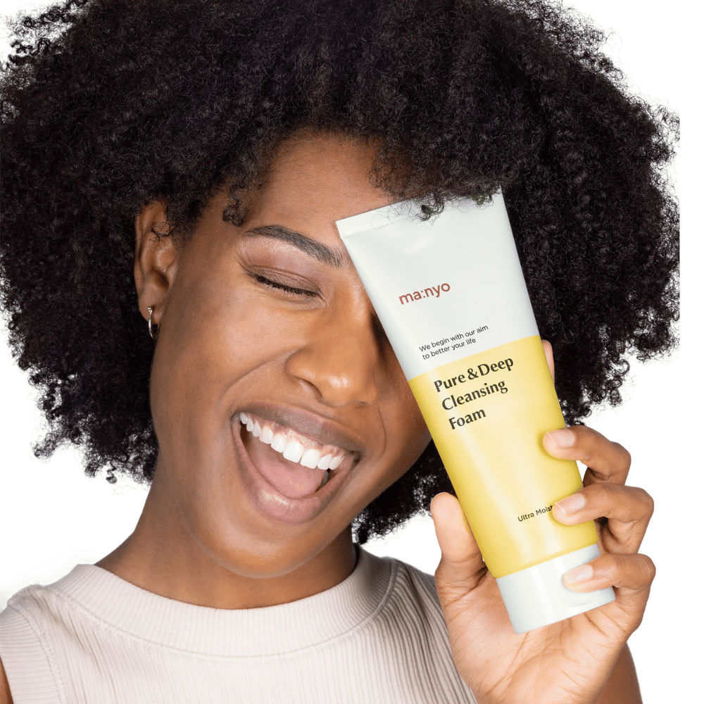 Ženska s kodrastimi lasmi drži Ma:nyo Pure & Deep Cleansing Foam – nežen korejski čistilec, ki učinkovito odstranjuje umazanijo in ličila, ne da bi izsušil kožo.