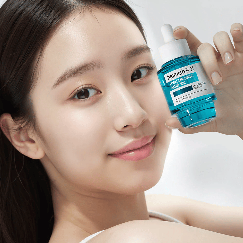 Nasmejana ženska s sijočo kožo drži stekleničko Heimish RX Hyaluronic Acid 2% Hydrating Serum, kar poudarja njegove vlažilne lastnosti.
