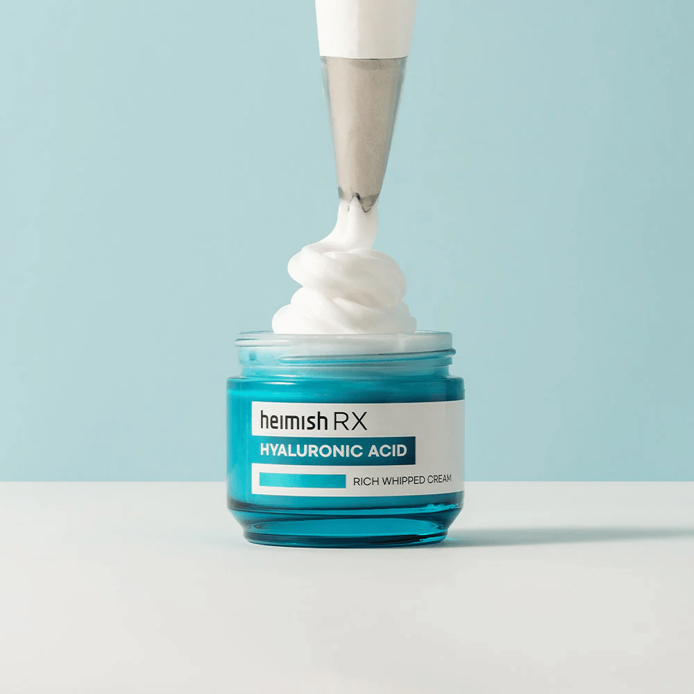 Heimish RX Hyaluronic Acid Rich Whipped Cream v modri stekleni posodi, kjer slaščičarska vrečka nanaša puhasto kremo. Vlažilna krema za obraz s hialuronsko kislino za intenzivno vlaženje in mehko, polno kožo.