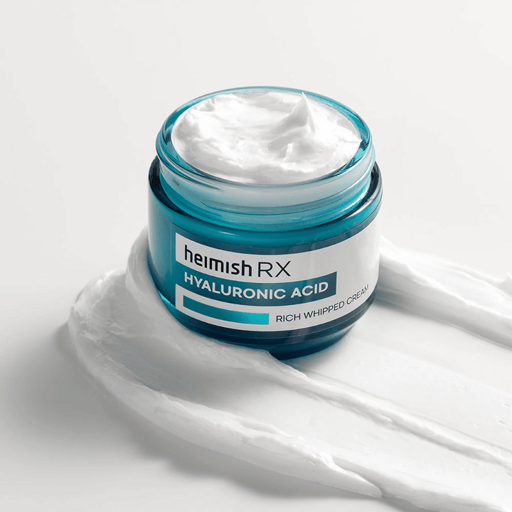 Heimish RX Hyaluronic Acid Rich Whipped Cream v modri stekleni posodi z bogato in puhasto konsistenco, razpršeno na beli podlagi. Intenzivno vlažilna krema za obraz s hialuronsko kislino za globoko vlaženje in mehko kožo.