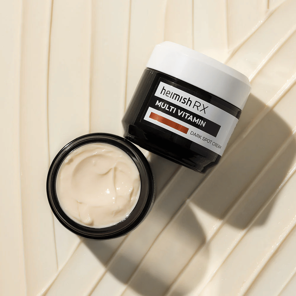 Heimish RX Multi Vitamin Dark Spot Cream – hranilna obrazna krema za zmanjšanje temnih madežev in izenačevanje tena kože. Kremasta tekstura kreme je prikazana v odprti posodi z elegantno embalažo.