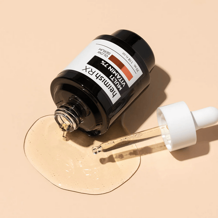Heimish RX Multi Vitamin 7% Glow Serum leži na bež ozadju s serumom razpršenim okoli stekleničke in pipeto napolnjeno z izdelkom, kar poudarja njegovo hranilno formulo.