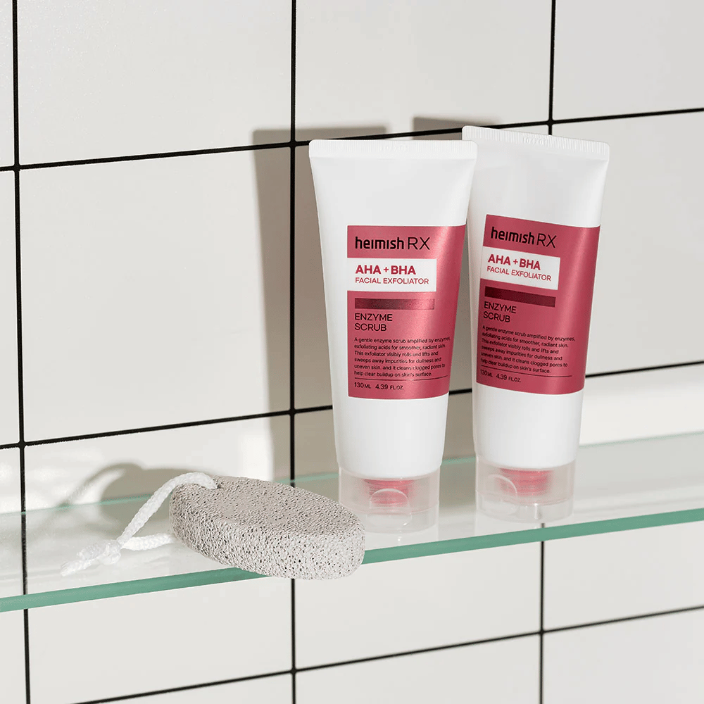 Dve tubi Heimish RX AHA + BHA Facial Exfoliator postavljeni na stekleno polico v kopalnici z belo ploščato steno. Poleg izdelkov leži plovec z belo vrvico. Elegantna in minimalistična slika nege kože.